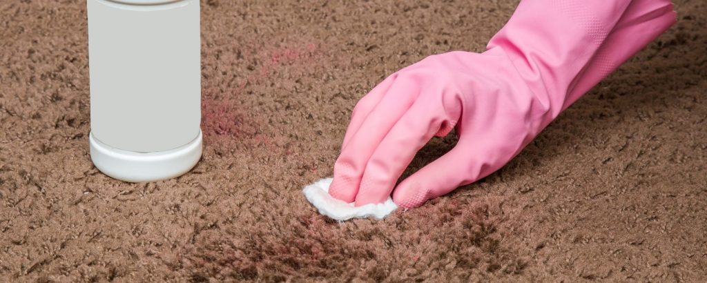 A pink gloved hand blotting a stain on brown carpet.